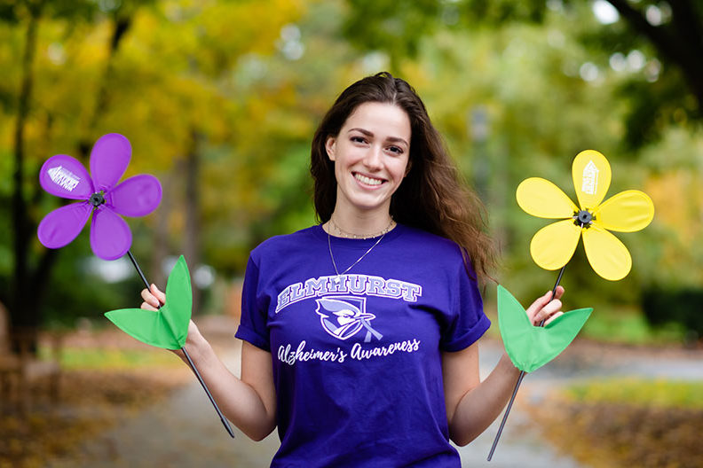 A female Elmhurst College student holds a colorful flower in each hand to promote Alzheimer's awareness month.